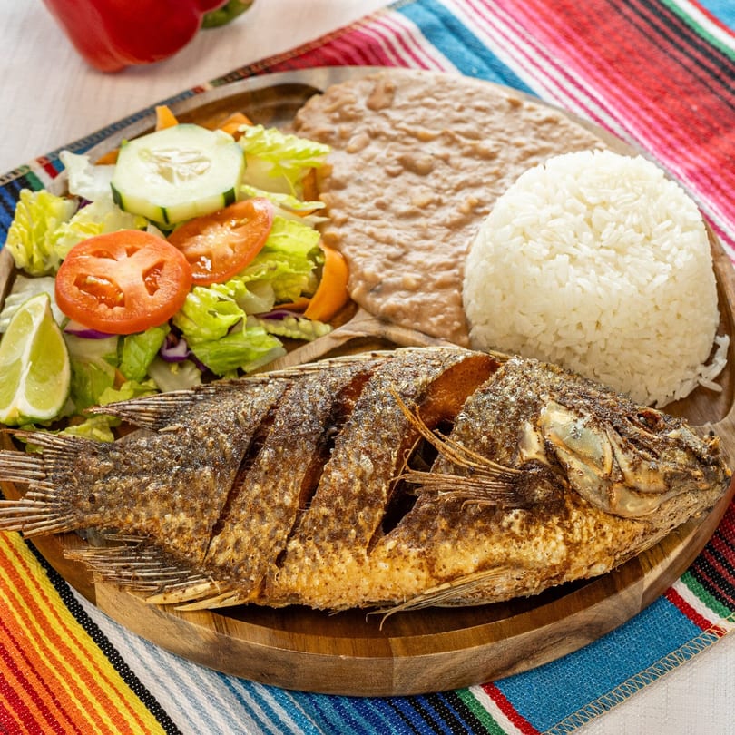 Fried whole fish with rice, refried beans, and a fresh salad on a wooden plate.
