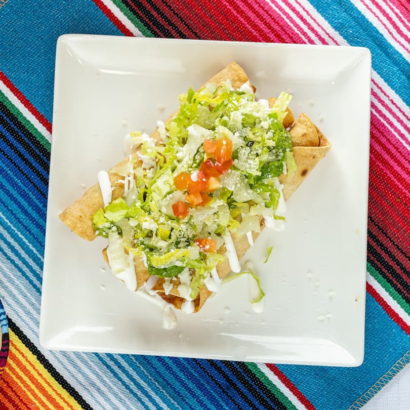 Crispy taquitos topped with shredded lettuce, diced tomatoes, crumbled cheese, and crema on a white plate.