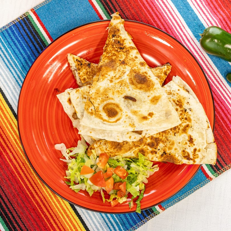 Golden-brown quesadillas with a side of lettuce and diced tomatoes on a red plate, atop a colorful striped placemat.