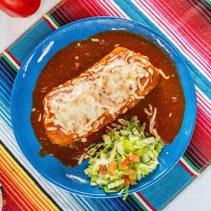A cheesy burrito smothered in red sauce, with a side of lettuce and diced tomatoes on a blue plate.