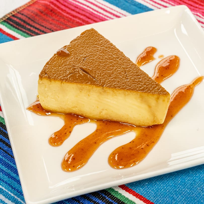 A slice of flan with caramel sauce on a white rectangular plate, on a colorful striped tablecloth.