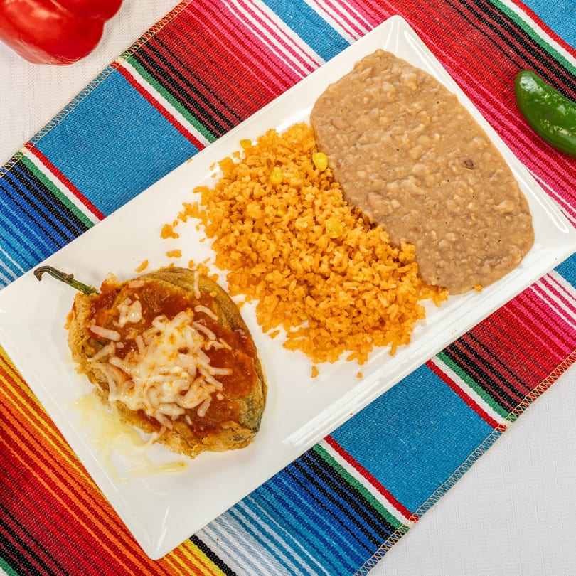Chili relleno with melted cheese, rice, and refried beans on a white plate over a colorful striped placemat.