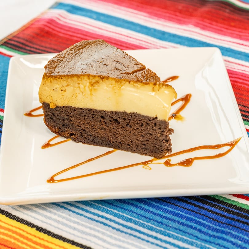 Slice of chocolate flan cake drizzled with caramel on a white plate, on a colorful striped tablecloth.