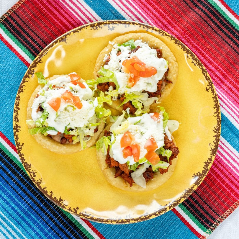 Three sopes with meat, lettuce, cheese, sour cream, and diced tomatoes on a yellow plate.