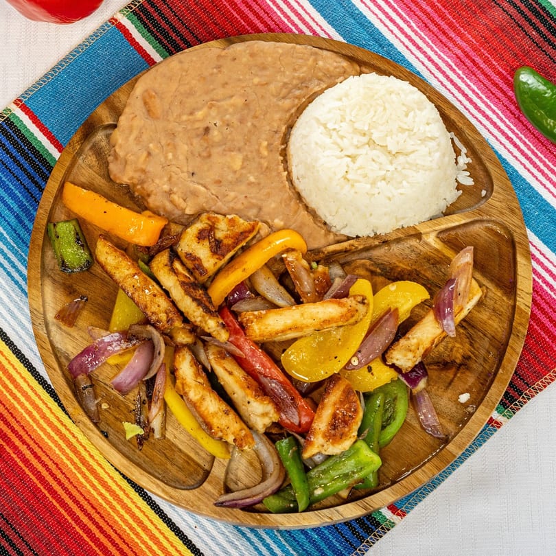 Chicken fajitas with colorful bell peppers and onions, served with rice and refried beans on a wooden plate.