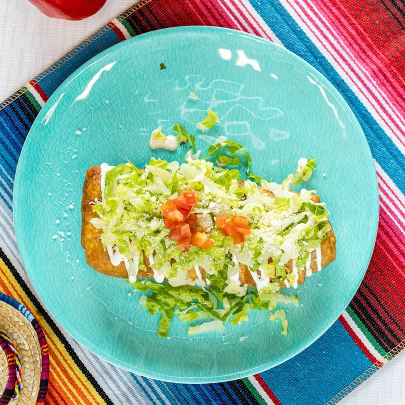 A golden-brown chimichanga topped with shredded lettuce, diced tomatoes, sour cream, and cheese on a blue plate.