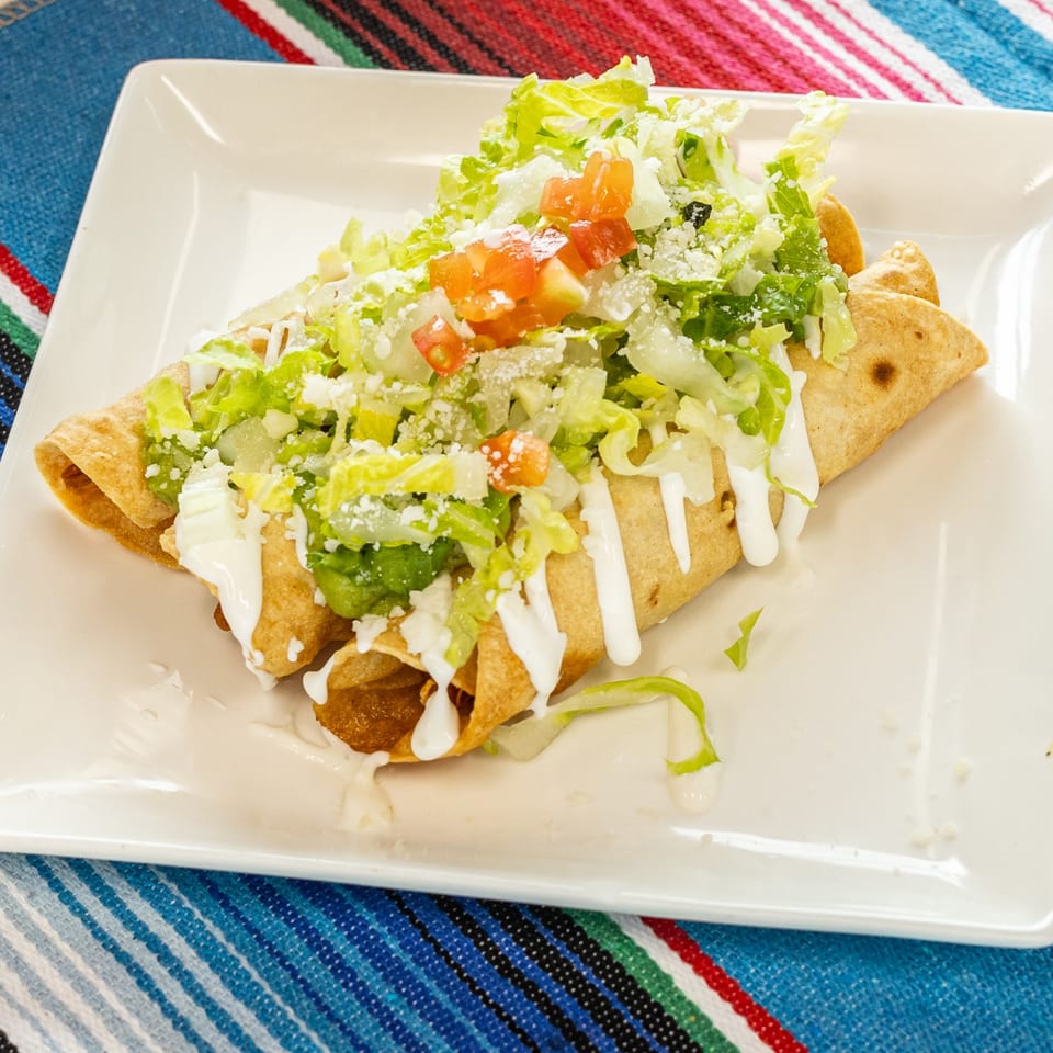 Two golden taquitos topped with shredded lettuce, diced tomatoes, crumbled cheese, and crema on a white plate.