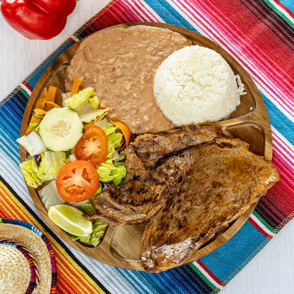 Grilled steak, refried beans, white rice, and a fresh salad with lime on a wooden plate.