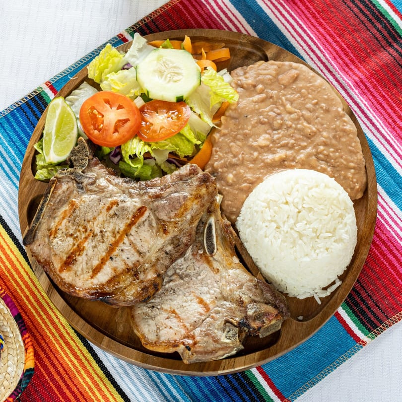 Grilled pork chops with rice, refried beans, and a fresh salad on a wooden plate.