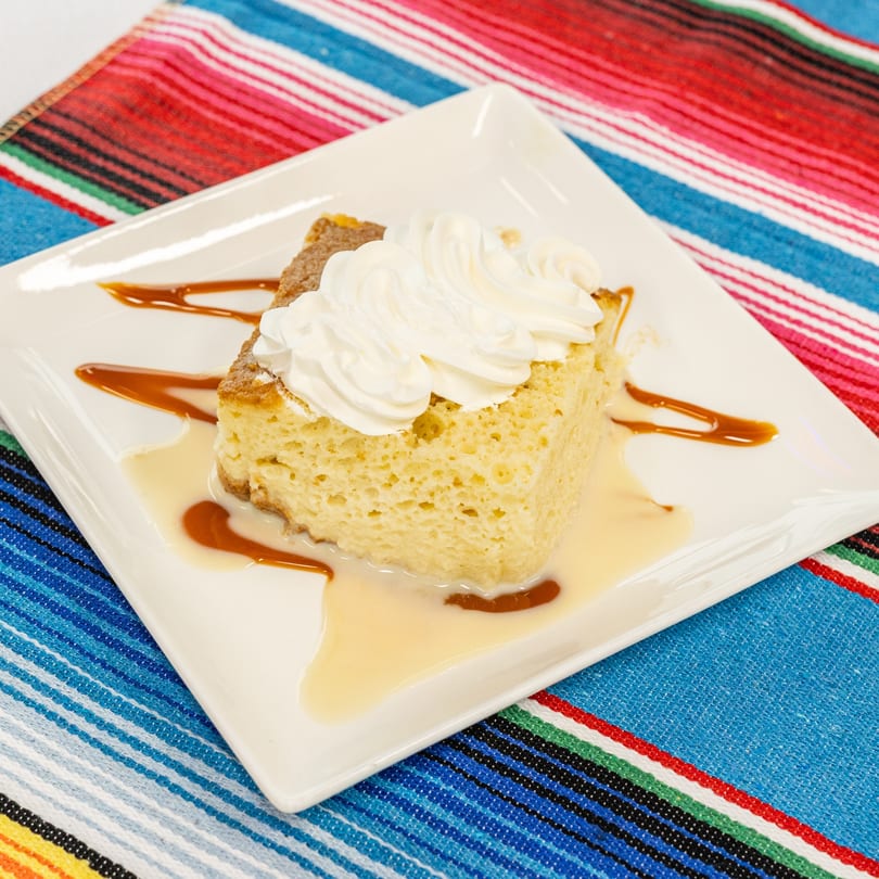 A slice of tres leches cake with whipped cream and caramel drizzle on a white plate.